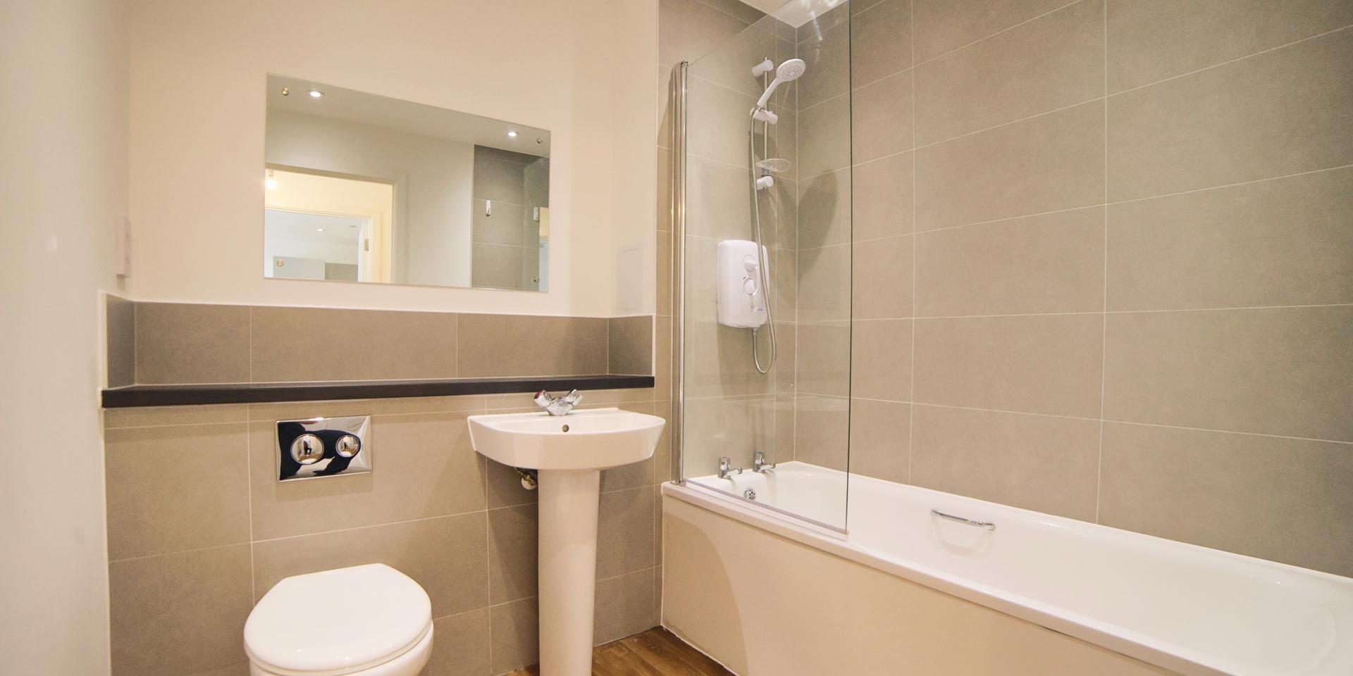 There is a white bathroom suite with a built-in shelf behind the sink and toilet. There is a glass shower screen and shower over the bath.