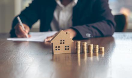Someone filling in a form and looking at piles of coins, next to the piles of coins there is a small model of a house.