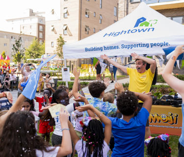 The entertainment at the event waving and getting children to do the same. They are in the sun, there are a lot of children in the photo, and a bouncy castle in the background