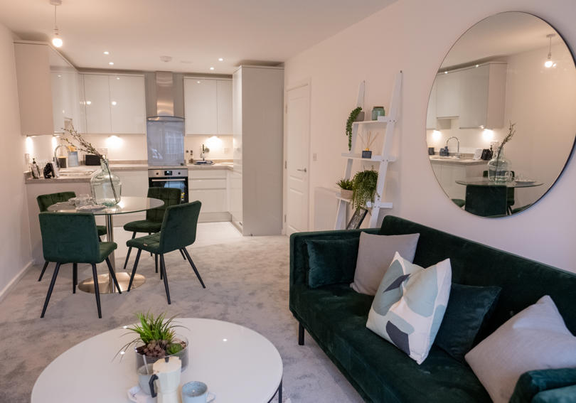 Inside of a new build flat. Emerald green velvet sofa and chairs, grey carpet and white kitchen units.