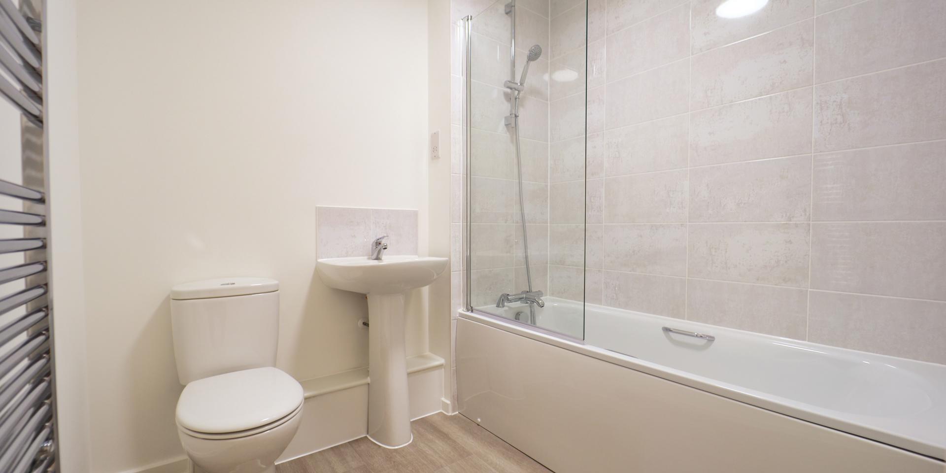 The bathroom consists of a white suite with a shower over the bath, glass shower screen, chrome heated towel rail and pedestal sink and toilet